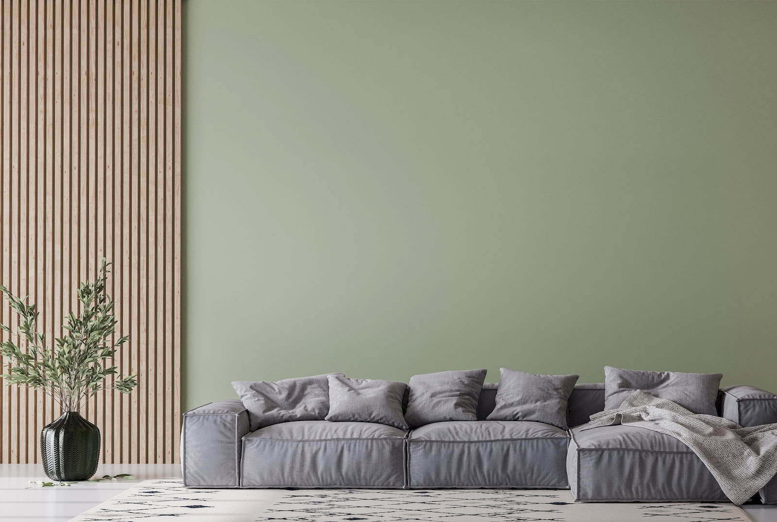 A modern living room with a soft sage-green painted wall, a grey modular sofa with cushions, a patterned rug, and a tall glass vase with leafy branches beside vertical timber slats.
